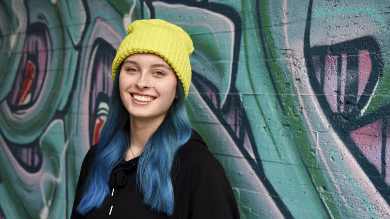 A young woman stands wearing a yellow beanie against a colorful wall.