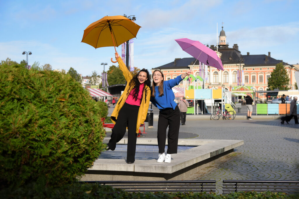 Two young women are holding umbrellas high in the air on a sunny square.